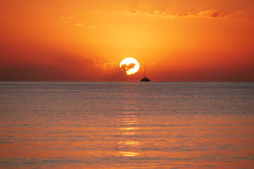 A breathtaking sunset over the calm sea with a sailboat on the horizon. Warm orange and golden tones fill the sky and reflect on the water. Travel, nature, relaxation, and maritime concept