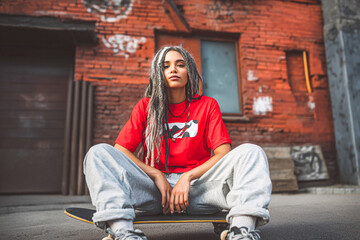 Woman with dreadlocks sitting on a skateboard in front of a brick building on a sunny day outdoors