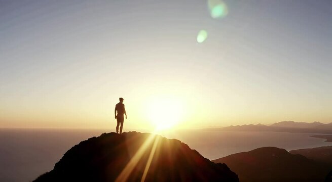 A lone man stands silhouetted against a vibrant sunset on a mountain peak overlooking the ocean