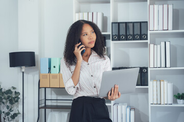 Businesswoman on a Call: A focused businesswoman stands in a modern office, holding a laptop and...