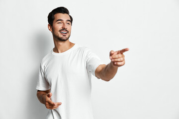 Smiling man in casual white t-shirt pointing with hand, looking at camera with cheerful expression,...