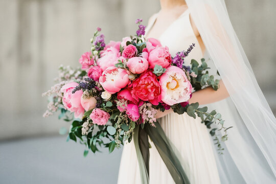 Bright and lush bridal bouquet of pink peonies, roses, and mixed florals with greenery, held by a bride in an elegant white gown