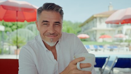Mature man drinking coffee by pool at luxurious resort on peaceful morning, enjoying relaxation. Portrait of happy mid adult, middle aged male on summer vacation, smiling.