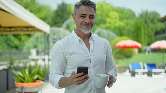 Portrait of happy mid adult man drinking morning coffee, holding phone on terrace at home. Confident mature male outdoor in garden, smiling. Businessman preparing to a usual work day.
