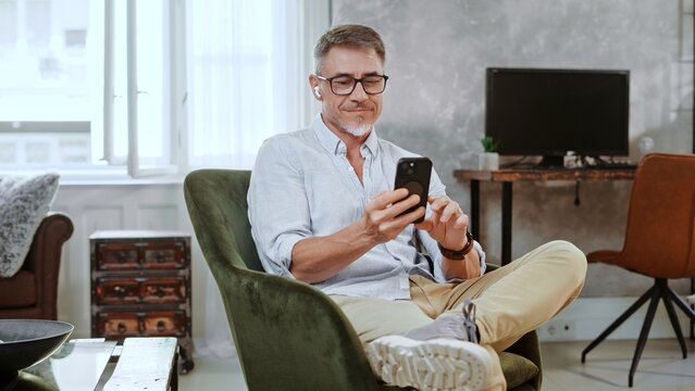 Entrepreneur relaxing in comfortable chair, working in home office. Mid adult man using smartphone and wearing earbuds. Happy, confident middle age male smiling.