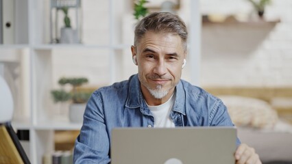 Portrait of happy mature man sitting at desk in white apartment, smiling. Confident businessman...