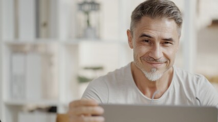 Portrait of happy mature man sitting at desk in white apartment, smiling. Confident businessman working on laptop in home office. Freelancer entrepreneur in remote work. Casual male on video call.