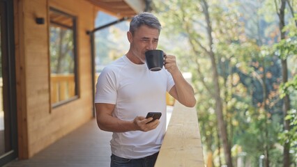 Senior man with grey hair reading messages on smartphone and drinking coffee on balcony of wooden chalet. Businessman on vacation in mountains. Relaxed lifestyle and serenity.