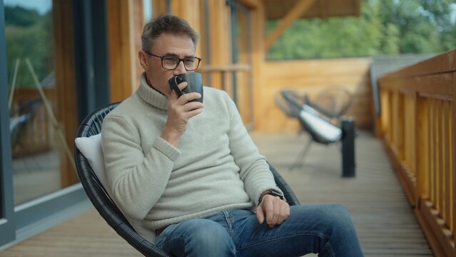 Mid adult man in 50s drinking hot coffee or tea from mug sitting on patio of contemporary wooden house, enjoying peaceful moment in nature. Serenity and tranquility, relaxed ideal lifestyle.