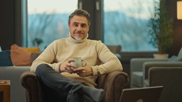 Portrait of confident middle aged man sitting at home, wearing turtleneck pullover. Mid adult older male in cosy living room, happy, smiling.