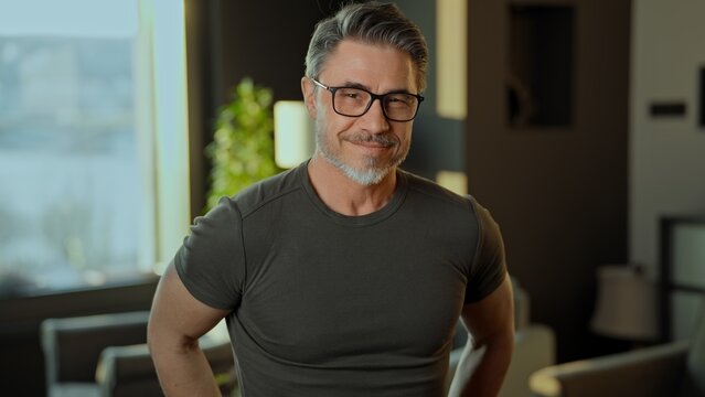 Portrait of a stylish mature man with glasses smiling in a contemporary interior at home. Happy confident fit muscular mid adult older male in 50s with friendly smile.  . - Powered by Adobe