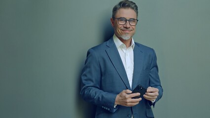 Executive corporate businessman in 50s standing against neutral background, dressed in formal business suit and glasses, holding smartphone. Portrait of happy middle aged confident man smiling.