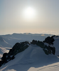 Sunrise over snowy mountain peaks with dramatic rock formations in the foreground. A serene alpine landscape bathed in soft winter light.