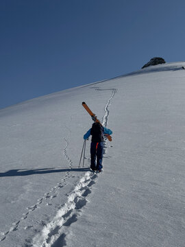 Lone skier ascending a pristine snowy slope under a deep blue sky, carrying skis and poles. A moment of solitude and alpine adventure.