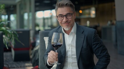 Elegant mature businessman smiling and holding glass of red wine, enjoying a moment of relaxation after work. Portrait of happy older man in restaurant, smiling, relaxing.