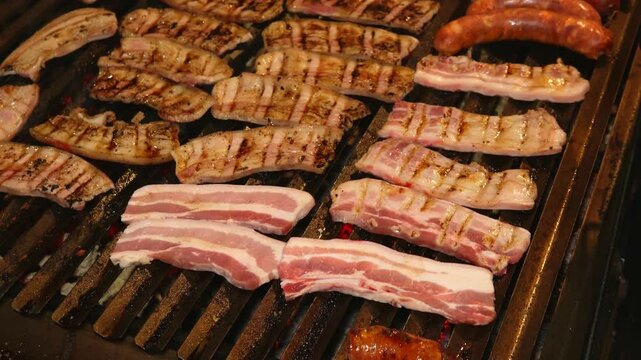 A close-up of a hot stainless steel barbecue grill with V-shaped grates. Sausages and pancetta sizzle on top, and fat drips down, smoking on the hot red coals. A bright, appetizing grilling scene.
