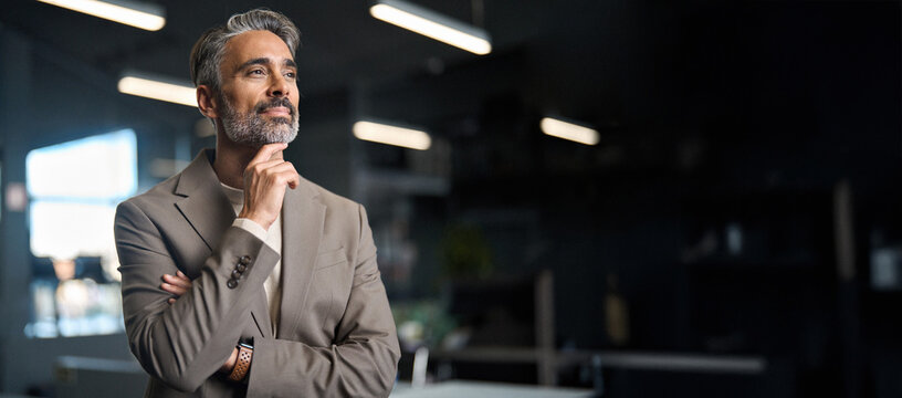 Middle aged business man investor, doubtful mature male ceo executive wearing suit standing in office holding hand in chin looking away at work thinking on leadership question, feeling doubt.