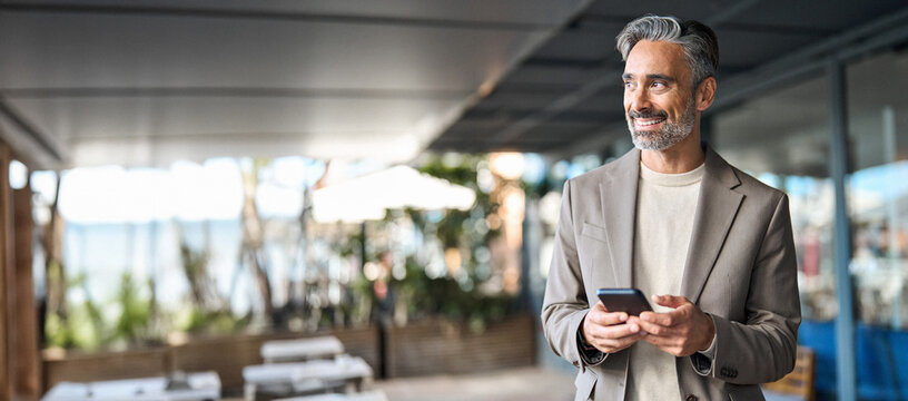 Happy mature business man executive using mobile cell phone corporate tech outdoors. Smiling handsome middle aged professional businessman holding cellphone looking away standing outside. Copy space - Powered by Adobe