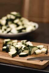Sliced zucchini on a wooden cutting board, with a knife next to it, and in the background a blurred bowl with more sliced vegetables, ready to cook
