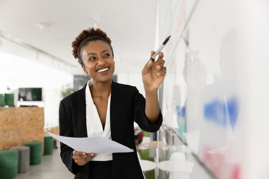 Smiling African woman leading brainstorming session, writing ideas on whiteboard, gives presentation or explaining concept visually, teaching class or workshop, planning project and strategy in office