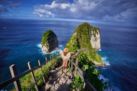 Happy woman traveler stands on steps of stairs leading to scenic Kelingking beach during a vacation to Nusa Penida Island in Indonesia, explore and travel to touristic beautiful destination