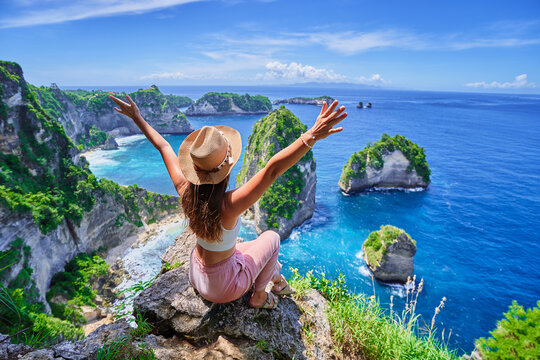 Woman traveler with open arms sitting on rock cliff and enjoying of scenic landscape of Diamond beach during vacation to Nusa Penida Island in Indonesia, travel to touristic beautiful destinations