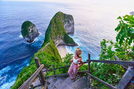 Woman traveler stands on steps of stairs and enjoying landscape of scenic Kelingking beach during vacation to Nusa Penida Island in Indonesia, travel to touristic beautiful destination