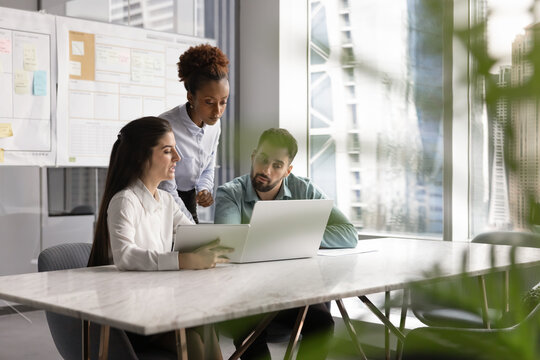 Three diverse focused colleagues take part in briefing using laptop and digital tablet, reviewing presentation or project update, collaborating on report, proposal, or marketing plan, analyzing data