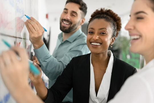 Happy African woman look at camera stand next to white board with two Latin colleagues generating ideas and jotting them down, working on plan or project together, engaged in successful collaboration - Powered by Adobe