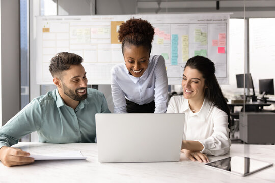 Three cheerful creative professionals, marketing department members gathered in office working together using laptop, brainstorming insights, share ideas for new campaign, service or product. Teamwork