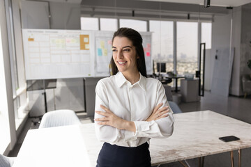 Optimistic Latina businesswoman standing in modern conference room, looking out window with...