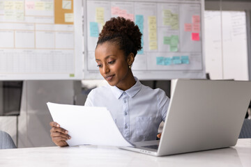 Young 25s African businesswoman working in office, sitting at desk with laptop, holding sheet, reviewing financial report or document, analyze data presented on paper, paying bills using e-bank system