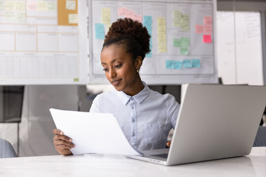 Young 25s African businesswoman working in office, sitting at desk with laptop, holding sheet, reviewing financial report or document, analyze data presented on paper, paying bills using e-bank system
