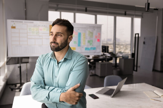 Thoughtful Hispanic professional man dressed in casual shirt standing alone with arms crossed in modern office, contemplating, looking out window, busy in brainstorming or planning, decision-making
