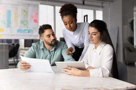 Three colleagues gathered around conference table, looking at tablet, hold document, reviewing information, discussing ideas while working on joint project together using digital and print resources