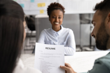 Smiling African woman passing job interview seated at desk in front of two HR managers hold resume...