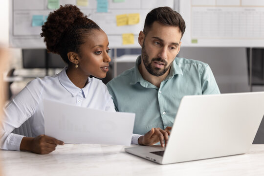 Two colleagues working together on document, cross-checking agreement details with information on laptop, preparing business proposal or client presentation, verifying financial data, invoices, bills