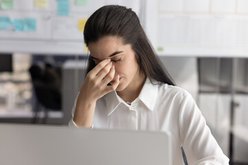 Tired woman sits at desk with laptop, closing dry or irritated eyes, rub nose bridge to reduce...