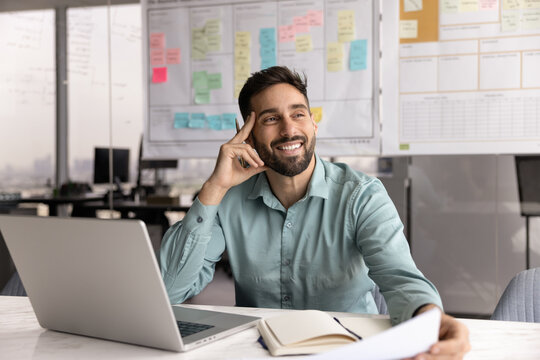 Pensive professional man sits in workplace desk with laptop, organizer and papers, looking into distance with joyful expression, thinks creatively or brainstorming ideas feeling inspired and motivated