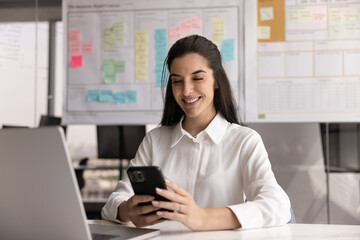 Professional woman sitting at desk using smartphone with contented smile, accessing work-related...