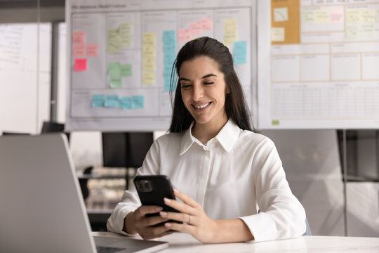 Professional woman sitting at desk using smartphone with contented smile, accessing work-related documents or files, managing tasks or to-do lists using mobile app, looking up information on internet