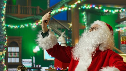 Fototapeta premium Professional Santa Claus taking phone selfie photos on Christmas set with large chairs, piles of presents, backdrops, stockings, fake fireplace, ornaments and other holiday props, camera A
