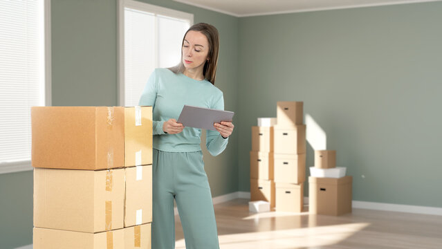 Woman with tablet checking moving boxes in empty room, organizing relocation or inventory process. - Relocation. Packing. Inventory management.
