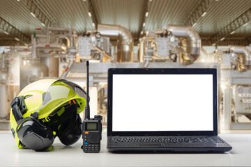 Laptop with blank screen, safety helmet, and walkie-talkie on table in industrial facility, symbolizing workplace communication and safety. - Industrial safety. Maintenance. Engineering.
