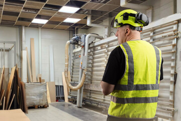 Worker in high-visibility vest and helmet inside a wood processing factory with stacked boards and...