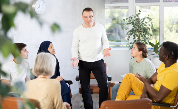 Smiling friendly man conducting informal work meeting with mixed age group of colleagues of different nationalities sitting on chairs in circle in office, discussing work-related situations