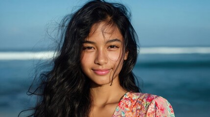 Portrait of a young woman with long dark hair. she is standing on a beach with the ocean in the background.