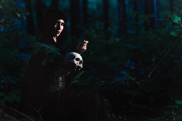Halloween theme. Gothic woman or witch in hood sitting in the forest with human skull and sickle or hook. Dramatic night image