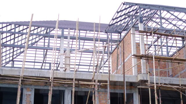 A building under construction with scaffolding and bamboo poles. The building is made of bricks and has a slanted roof
