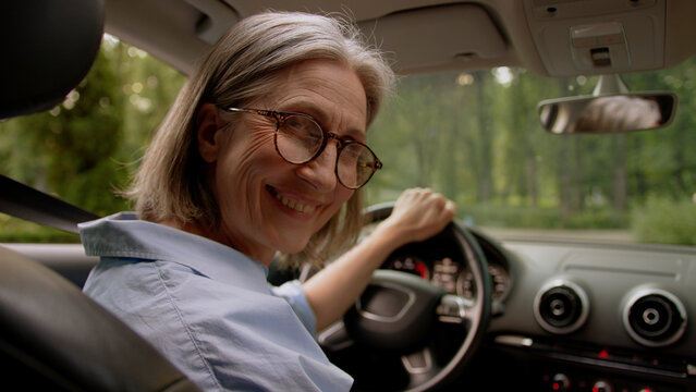 Back view happy smiling Caucasian aged elderly female cheerful mature senior woman taxi car driver riding in automobile driving auto steering wheel stop vehicle turning looking camera passenger smile